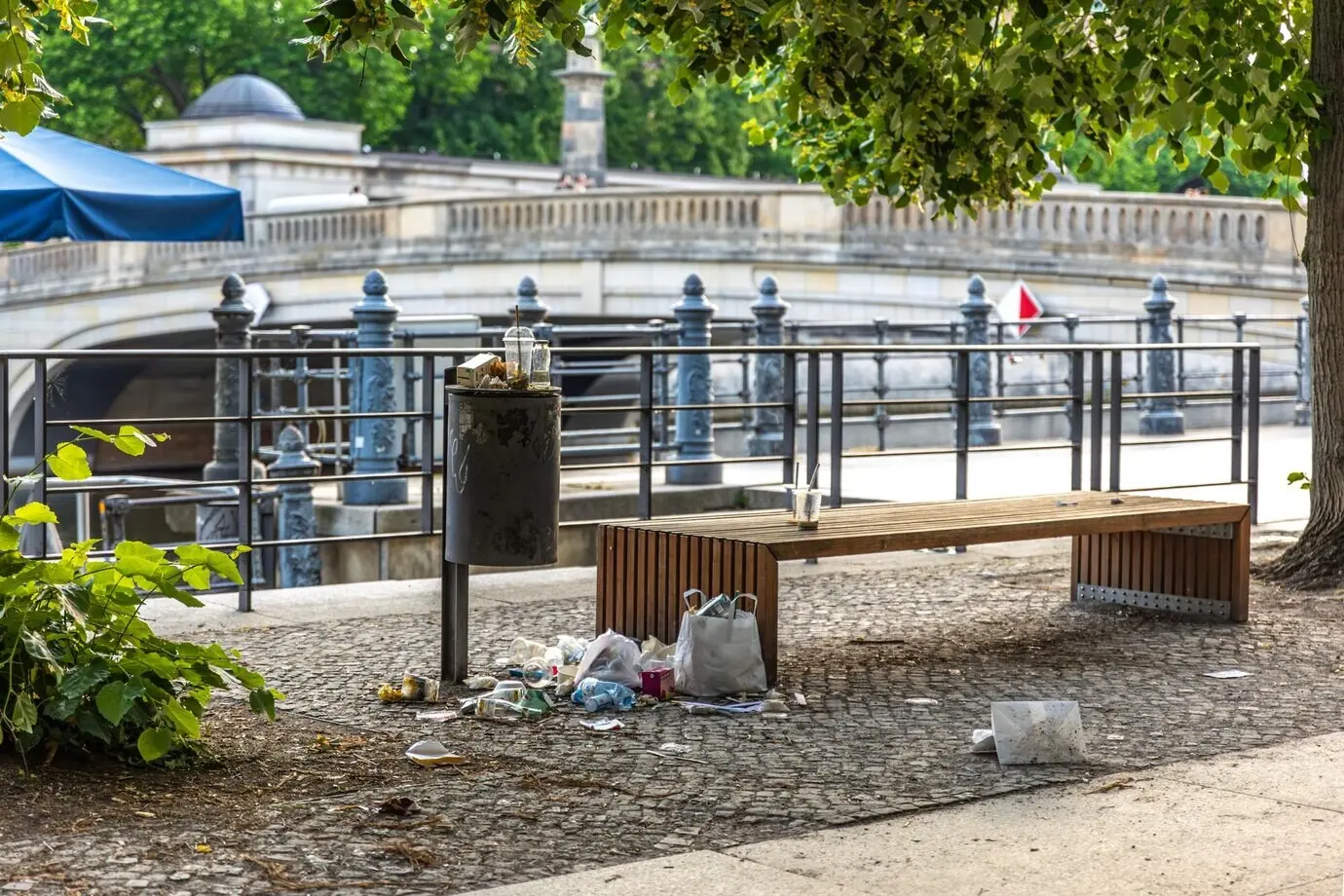 Überquellender Mülleimer neben einer Bank am Flussufer, städtisches Durcheinander und städtische Verschmutzung.